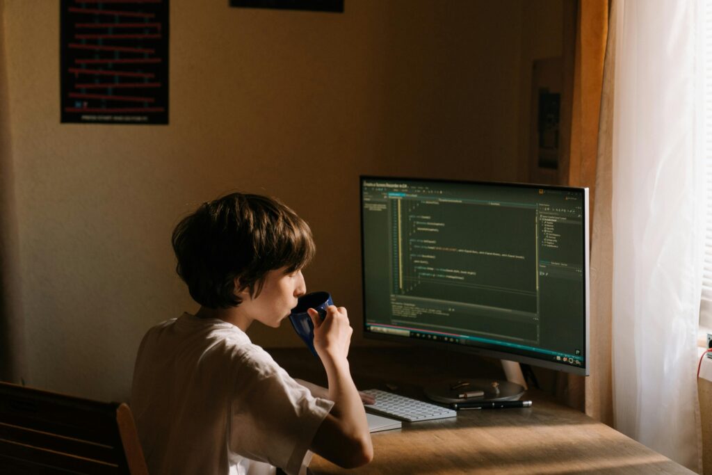 A young person coding at a desk with a computer and drinking from a mug.